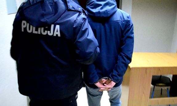 Policjant w kurtce z napisem Policja na plecach z zatrzymanym, który na rękach trzymanych z tyłu ma założone kajdanki. Widok z tyłu.