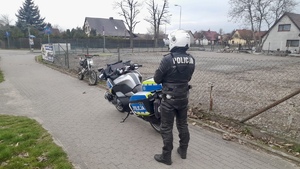 Policjant stoi przy motocyklu, w tle stoi motor opary o siatkę płotu.