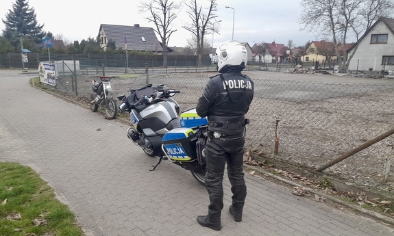 Policjant stoi przy motocyklu, w tle stoi motor opary o siatkę płotu.