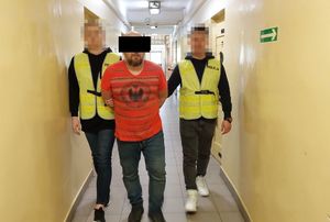 Dwaj policjanci w żółtych kamizelkach z napisem Policja prowadzą zatrzymanego mężczyznę korytarzem. Widok z przodu.
