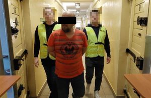 Dwaj policjanci w żółtych kamizelkach z napisem Policja prowadzą zatrzymanego mężczyznę korytarzem. Widok z przodu.