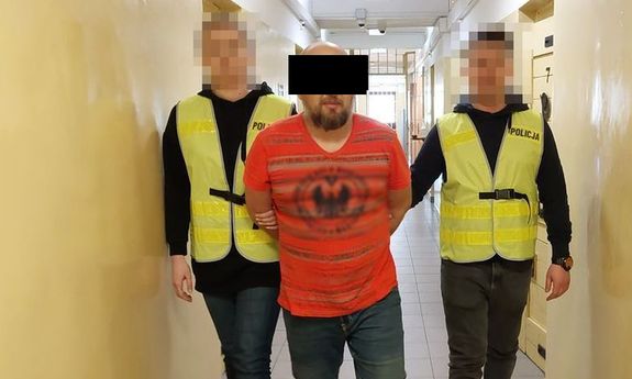 Dwaj policjanci w żółtych kamizelkach z napisem Policja prowadzą zatrzymanego mężczyznę korytarzem. Widok z przodu.