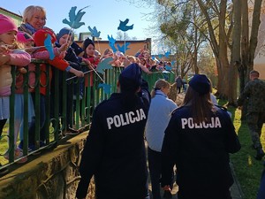 Idące dwie policjantki a przy barierce z boku widać dzieci z opiekunką, które machają niebieskimi ptakami z papieru.