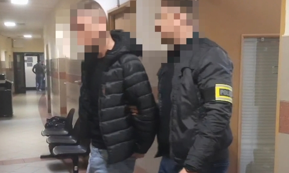 Policjant prowadzi zatrzymanego mężczyznę zakutego w kajdanki.