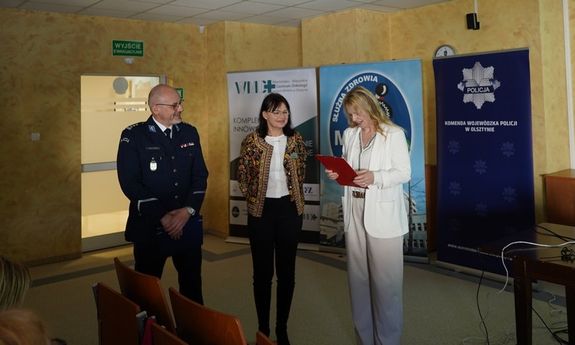 Umundurowany policjant i dwie kobiety stoją na środku przed osobami zebranymi na konferencji.