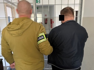 Nieumundurowany policjant z zatrzymanym mężczyzną. Przed nimi krata. Widok z tyłu.
