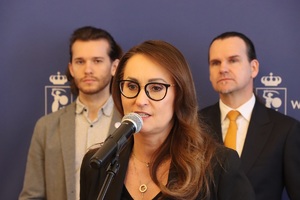 Na zdjęciu uczestnicy konferencji. Na pierwszym planie kobieta mówi do mikrofonu, w tle stoi dwóch mężczyzn.
