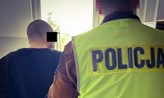 Zatrzymany prowadzony przez policjanta w żółtej kamizelce z napisem Policja na plecach.