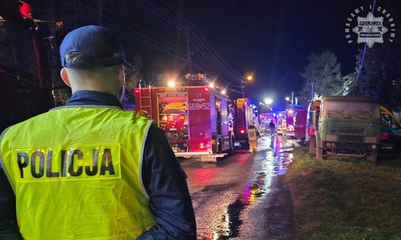 Policjant zabezpieczający miejsce zdarzenia stoi tyłem w żółtej kamizelce z napisem Policja. Przed nim widać wozy strażackie biorące udział w akcji gaszenia pożaru.
