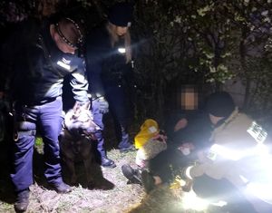 Przy odnalezionej leżącej na ziemi kobiecie kuca strażak, a z drugiej strony stoi policjantka i przewodnik z psem Tolo.