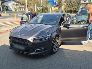 Dwaj ubrani po cywilnemu policjanci przy zaparkowanym fordzie z otwartymi drzwiami.