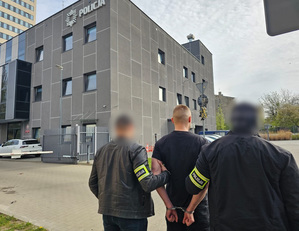 Dwaj funkcjonariusze prowadzą zatrzymanego w kajdankach.