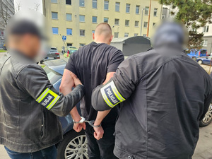 Dwaj funkcjonariusze prowadzą zatrzymanego w kajdankach.