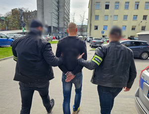 Dwaj funkcjonariusze prowadzą zatrzymanego w kajdankach.