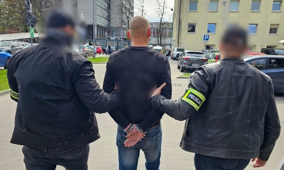 Dwaj funkcjonariusze prowadzą zatrzymanego w kajdankach.
