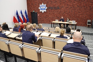 Na podeście, za stołem, w sali konferencyjnej siedzi nadinsp. dr Tomasz Michułka, Zastępca Komendanta Głównego Policji. Obok niego Pełnomocniczka Komendanta Głównego Policji ds. wdrażania Planu Równości Płci w Policji na lata 2023-2026. Na pierwszym planie uczestnicy spotkania.