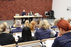 Na podeście, za stołem, w sali konferencyjnej siedzi nadinsp. dr Tomasz Michułka, Zastępca Komendanta Głównego Policji. Obok niego Pełnomocniczka Komendanta Głównego Policji ds. wdrażania Planu Równości Płci w Policji na lata 2023-2026. Na pierwszym planie uczestnicy spotkania.