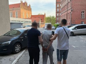 Dwaj ubrani po cywilnemu policjanci prowadzą zatrzymanego.