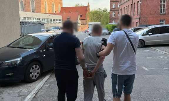 Dwaj ubrani po cywilnemu policjanci prowadzą zatrzymanego.