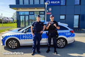 Dwaj funkcjonariusze stoją przy radiowozie zaparkowanym przed komendą Policji.