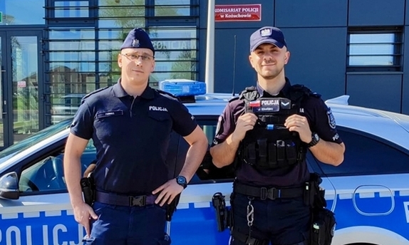 Dwaj funkcjonariusze stoją przy radiowozie zaparkowanym przed komendą Policji.