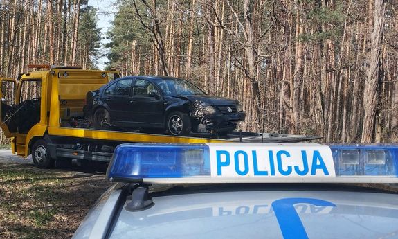 Na pierwszym planie widoczny dach radiowozu i belka z napisem Policja, z tyłu laweta a na niej rozbite auto.