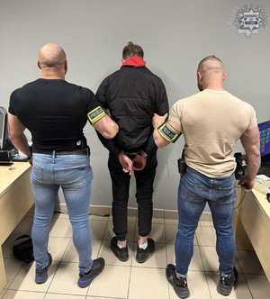 Dwaj nieumundurowani policjanci oraz zatrzymany mężczyzna z kajdankami założonymi na ręce z tyłu. Widok z tyłu.