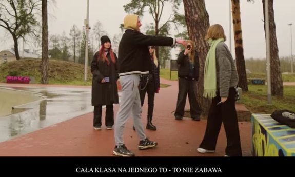 kadr z teledysku. chłopak pokazuje na dziewczynę i wytyka ją. w tle stoją inne dzieci.