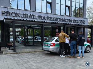Trzech funkcjonariuszy stoi z zatrzymanym mężczyzną przy radiowozie. W tle budynek Prokuratury Regionalnej w Łodzi.