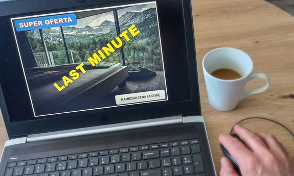 Na ekranie monitora wyświetla się zdjęcie z napisem Super oferta, Last minute, najniższa cena za dobę. Obok widoczny kubek kawy i dłoń na myszce komputera.