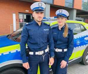 Dwoje umundurowanych policjantów ruchu drogowego na pierwszym planie, za nimi widać policyjny radiowóz.