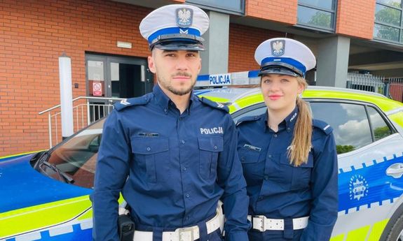 Dwoje umundurowanych policjantów ruchu drogowego na pierwszym planie, za nimi widać policyjny radiowóz.