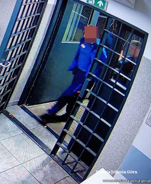 Kadr z monitoringu. Zatrzymany mężczyzna prowadzony przez policjanta przez okratowane drzwi.