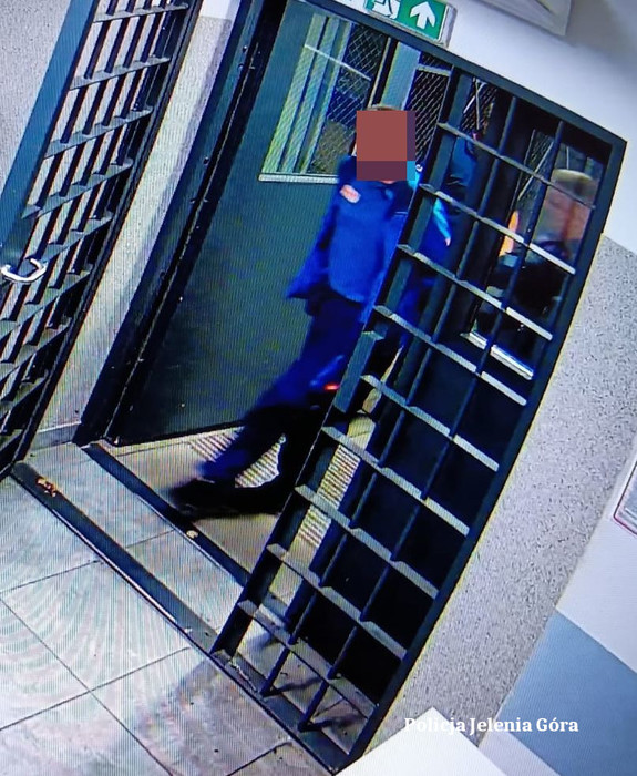 Kadr z monitoringu. Zatrzymany mężczyzna prowadzony przez policjanta przez okratowane drzwi.
