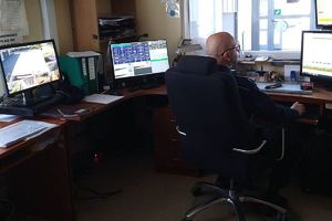 Zdjęcie przedstawia pomieszczenie policyjne - dyżurkę, gdzie znajdują się włączone komputery z monitorami i przy jednym z komputerów siedzi policjant.