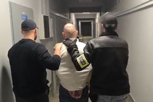 Na zdjęciu dwóch nieumundurowanych policjantów, z których jeden ma założoną na ręku żółtą opaskę z napisem POLICJA, i pośrodku nich zatrzymany mężczyzna. Policjanci prowadzą go korytarzem.