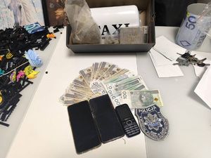 Na zdjęciu leżące na białym podłożu papierowe banknoty, trzy telefony komórkowe i obok nich naszywka policyjna wydziału patrolowo-wywiadowczego Komendy Miejskiej Policji w Łodzi. Na zdjęciu widoczne są też klucze, puszka, plastikowe elementy i karton, w którym znajdują się dwa worki z zawartością oraz inne przedmioty.