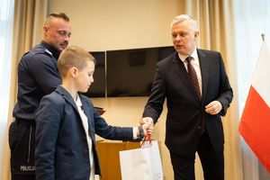 Mężczyzna w stroju cywilnym wręcza chłopcu białą torbę. Obok stoi policjant.