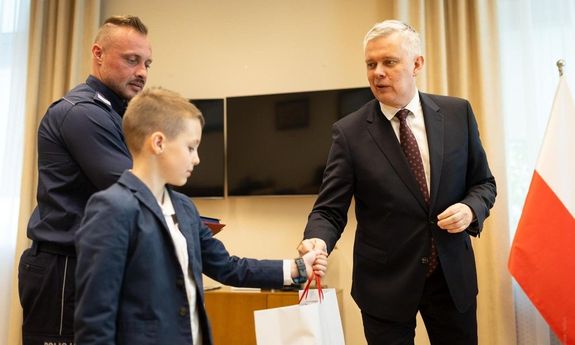 Mężczyzna w stroju cywilnym wręcza chłopcu białą torbę. Obok stoi policjant.