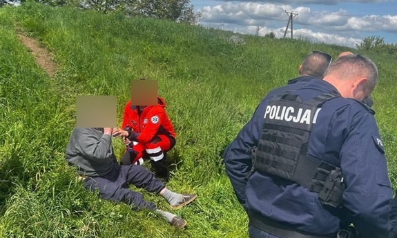 Ratownik medyczny podaje mężczyźnie siedzącemu na wzgórzu wodę obok policjanci.
