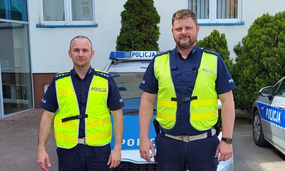 Dwaj policjanci stoją przed radiowozami zaparkowanymi przed budynkiem komendy.