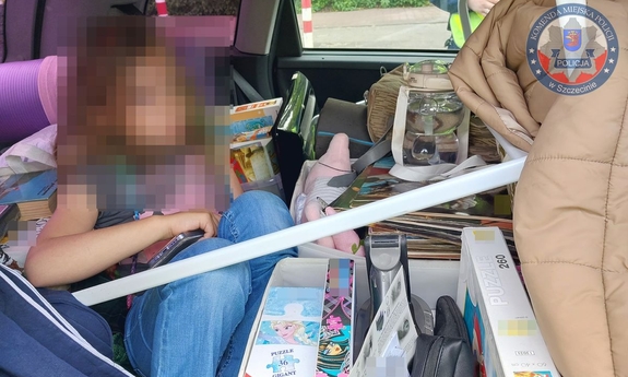 Na zdjęciu dziewczynka siedzi na tylnej kanapie samochodu Wokół dziecka znajdują się kartony, zabawki i inne rzeczy.