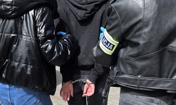 Dwie nieumundurowane policjantki prowadzą zatrzymaną, która na rękach trzymanych z tyłu ma założone kajdanki. Widok z tyłu.