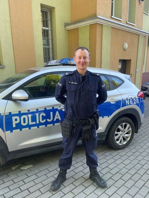 Umundurowany policjant stoi przed radiowozem i pozuje do zdjęcia.