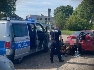 Policjant przy radiowozie. Obok na ziemi lezy zatrzymany mężczyzna, któremu policjant zakłada kajdanki.