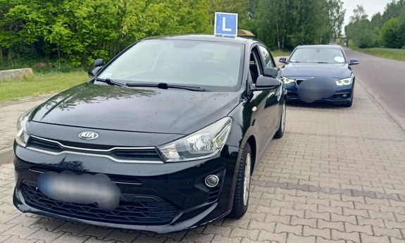 Na poboczu drogi stoi zaparkowane auto koloru czarnego z oznaczeniem na dachu literą L, za nim stoi granatowy samochód osobowy.