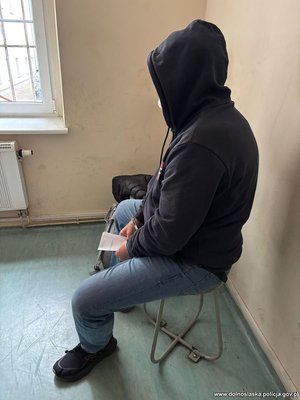 Zatrzymany mężczyzna siedzący w kajdankach na rękach i kapturze na głowie.
