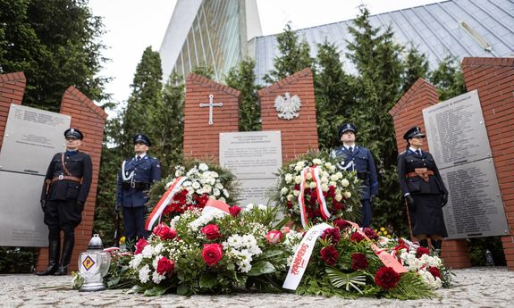 Wieńce i kwiaty przed ponikiem. Po bokach stoi czworo policjantów pełniących warte przy grobie.