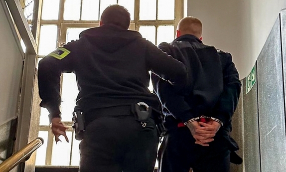 Policjant prowadzi schodami do góry zatrzymanego w kajdankach.