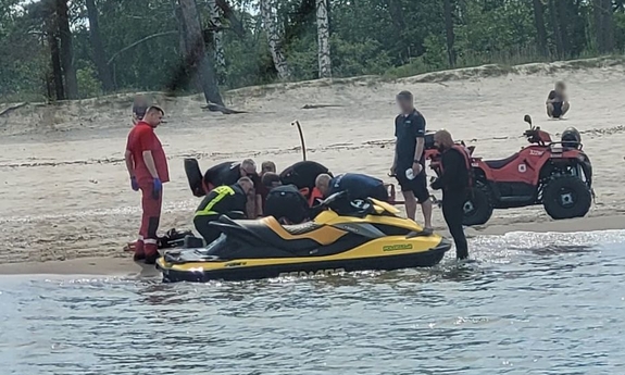 Skuter wodny przy brzegu. Obok quad i służby ratunkowe udzielające pomocy poszkodowanemu.